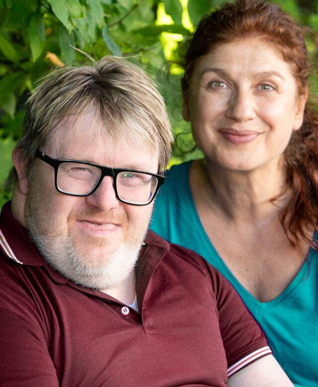 Un uomo con disabilità è seduto su una panchina con un’accompagnatrice; entrambi guardano la fotocamera con un’espressione cordiale.